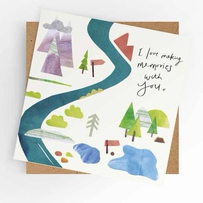 Square illustrated greeting card with a soft watercolour and paper collage landscape scene featuring a winding river path, mountains, trees, signpost, and red mailbox in teal, green, purple, red, and grey tones, with handwritten-style script reading "I love making memories with you," shown alongside a kraft brown envelope.